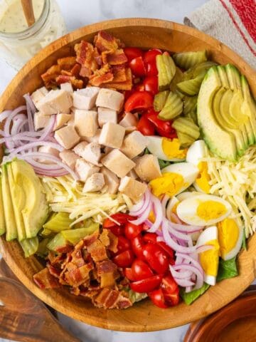 Loaded Chicken Bacon Club Sandwich Salad with Avocado salad filling a large wooden salad bowl. Wooden utensils, salad bowls and a jar of dressing around the bowl.