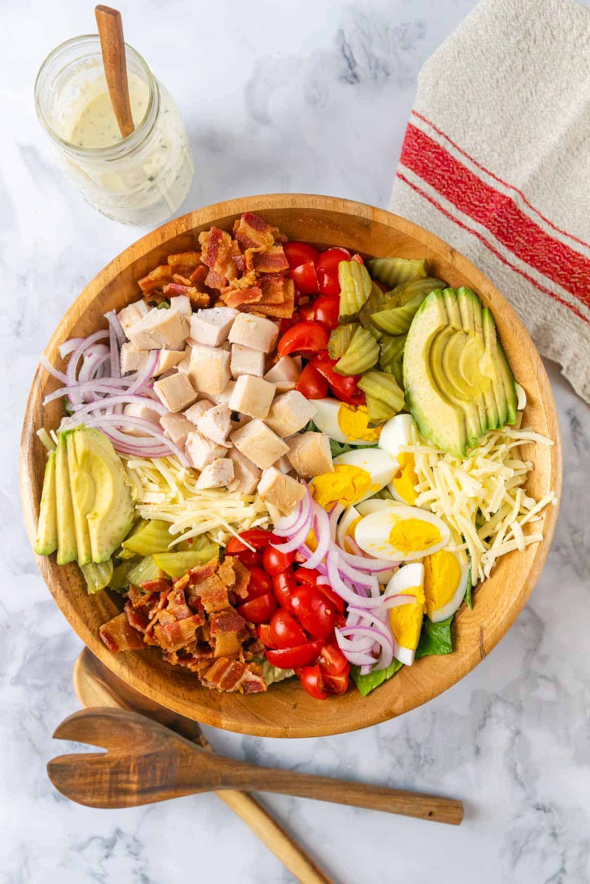 A large wooden salad bowl filled with club salad. Lettuce, bacon, chunks of chicken, avocado, eggs, cheese, pickles and red onion piled into the bowl. Wooden salad tongs near the bowl. A linen napkin and mason jar of creamy dressing in the top corner.