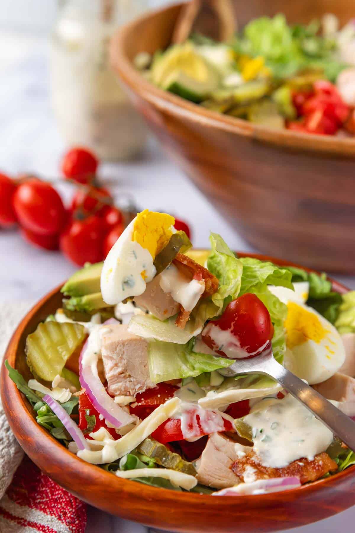 A small wooden bowl of club salad with dressing drizzled over the top. A fork with salad hovering above the bowl. In the foreground is a beige and red linen napkin. In the background is a large wooden salad bowl, a jar of dressing and tomatoes in the vine.