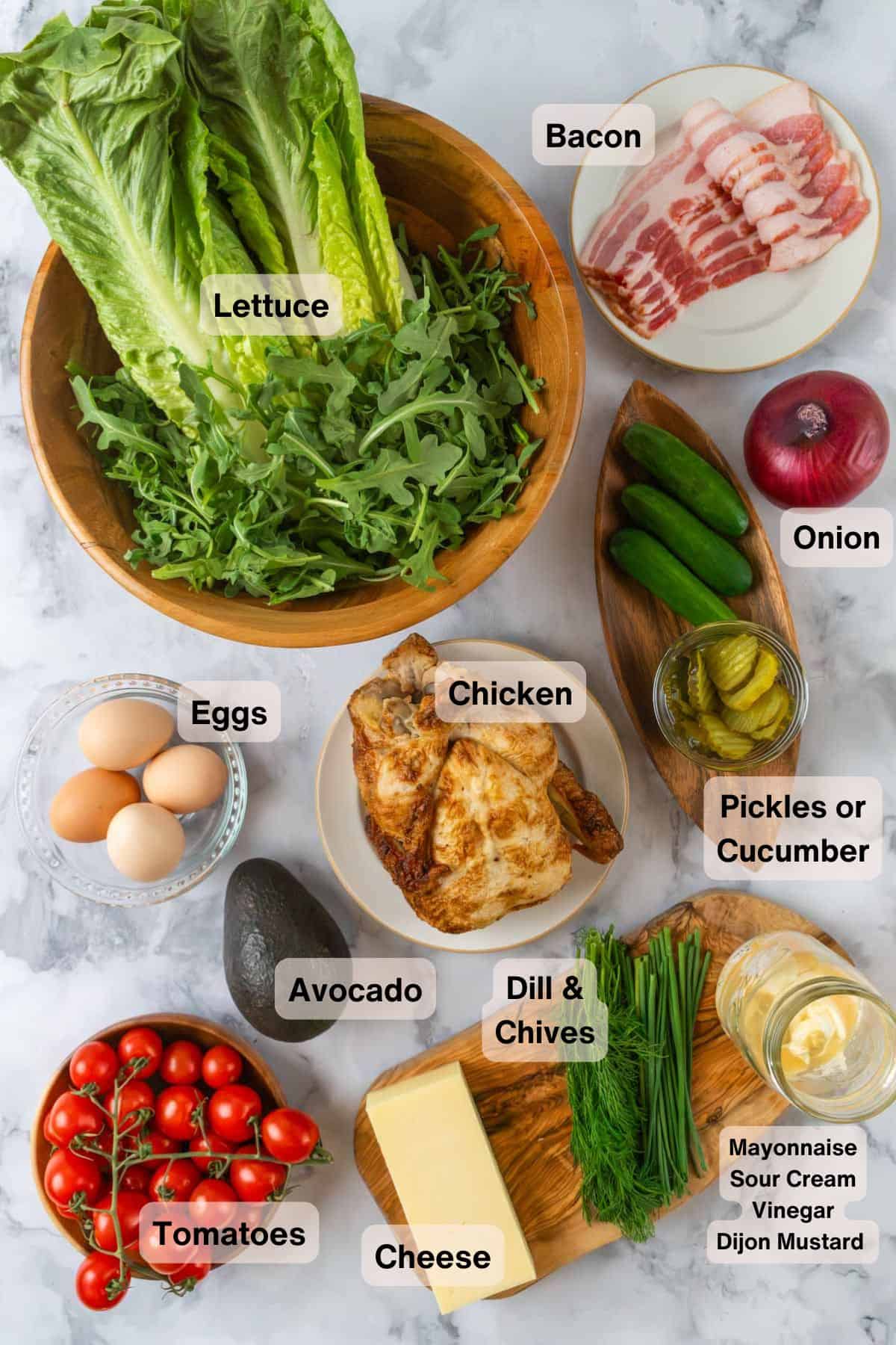 Ingredients for a Loaded Chicken Bacon Club Sandwich Salad with Avocado scattered around a white marble countertop in various bowls and dishes.