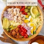 Loaded Chicken Bacon Club Sandwich Salad with Avocado filling a large wooden salad bowl with small bowls, a jar of dressing, tomatoes and wooden salad serving utensils to the side on a white marble countertop.
