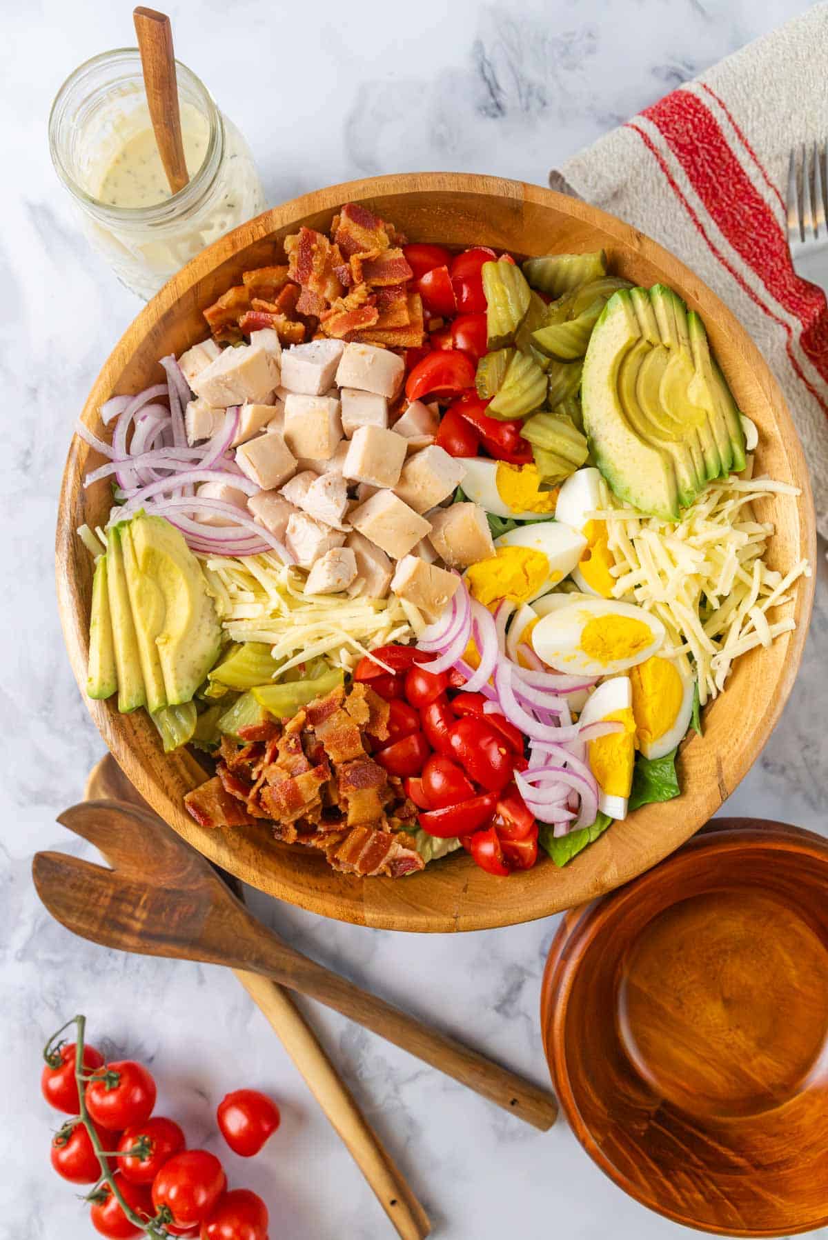 A large wooden salad bowl filled with club salad. Lettuce, bacon, chunks of chicken, avocado, eggs, cheese, pickles and red onion piled into the bowl. Small serving bowls and wooden salad tongs near the bowl. A fork, linen napkin and mason jar of creamy dressing in the top corners.