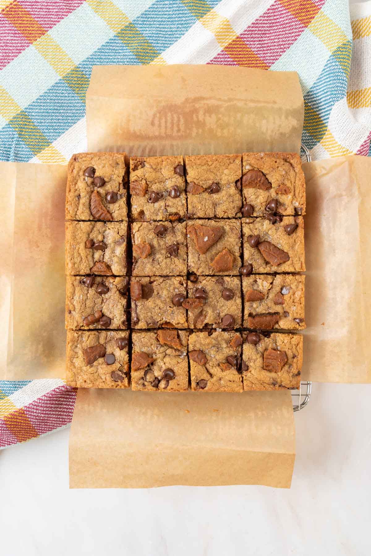 Chewy Biscoff blondies baked golden brown and cut into squares on parchment paper placed on a wire cooling rack on top of a cholorful checkered kitchen towel.