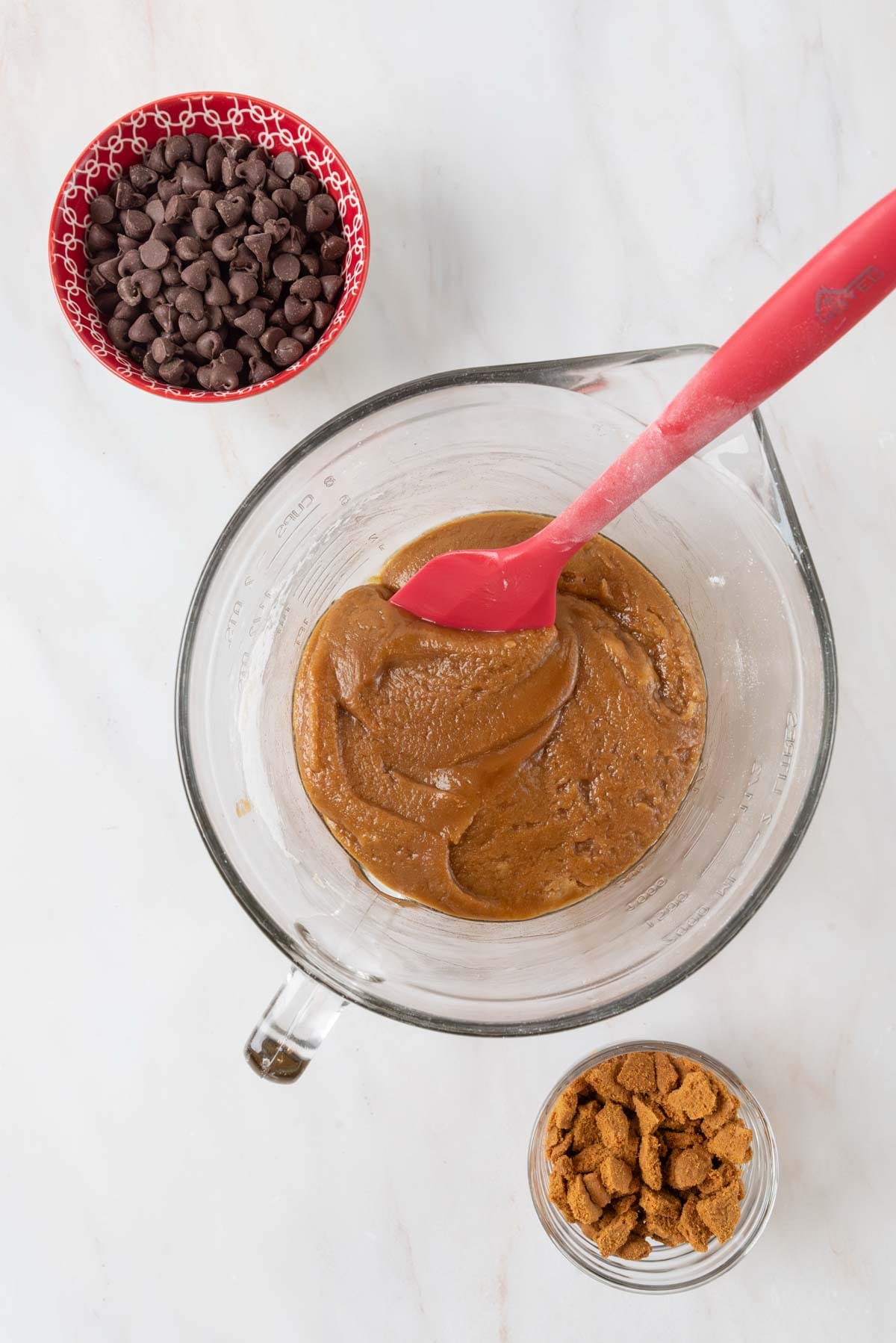 A large clear glass mixing bowl with blondie batter mixed together. Two bowls filled with chocolate chips and cookie crumbles sit beside the larger bowl.