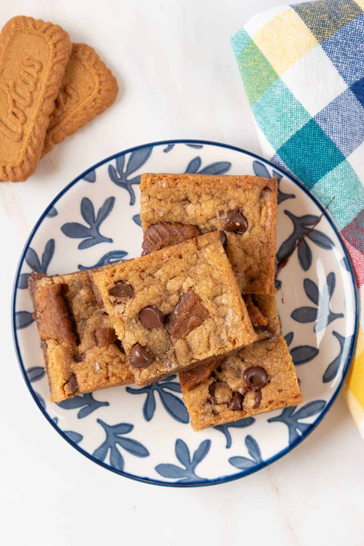 Four blondie squares resting on a blue and white dessert plate with a colorful checkered napkin in the corner and two Biscoff cookies on the countertop.