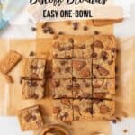 A wide shot of a pan of chewy Biscoff blondies cut into squares. The blondies are on a piece of parchment paper. They are spread apart a little bit. Chocolate chips and cookie crumbles are scattered around the countertop. A small bowl of Biscoff cookie spread with a spoon is at the bottom and a colorful towel at the top. Pinterest graphics overlayed.