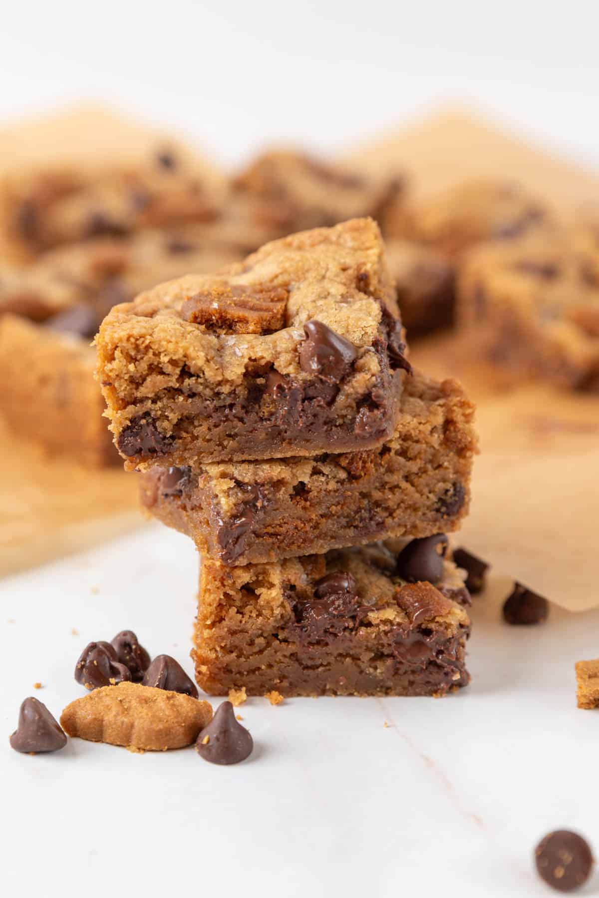 A closeup of a stack of three chewy brown butter Biscoff brownies baked golden brown. Chocolate chips and crumbled cookies are scattered in the foreground. More blondies are out of focus in the background.