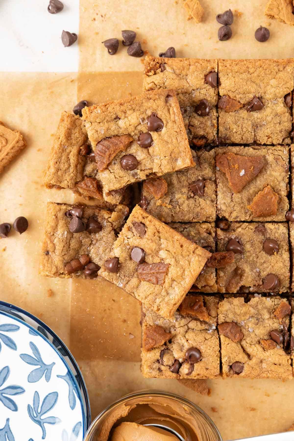 A closeup of chewy brown butter Biscoff brownies baked golden brown and cut into squares on a parchment paper. A stack of small blue and white dessert plates in the corner and a bowl of Biscoff cookie spread partially in the frame.