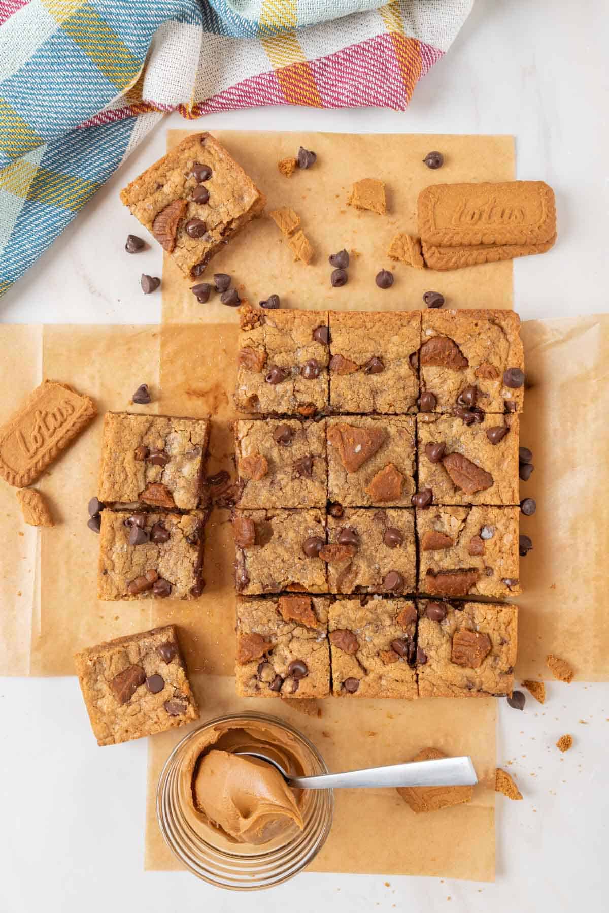 A wide shot of a pan of chewy Biscoff blondies cut into squares. The blondies are on a piece of parchment paper. They are spread apart a little bit. Chocolate chips and cookie crumbles are scattered around the countertop. A small bowl of Biscoff cookie spread with a spoon is at the bottom and a colorful towel at the top.