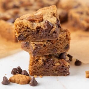 A closeup of a stack of three Chewy Biscoff Blondies on a white marble countertop with more out of focus in the background.