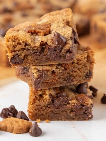 A closeup of a stack of three Chewy Biscoff Blondies on a white marble countertop with more out of focus in the background.