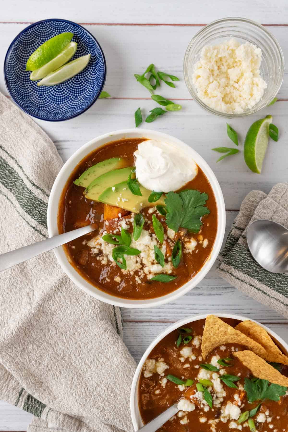 A white bowl filled with sweet potato black bean soup, garnished with queso fresco, sour cream, green onion, cilantro and avocado on a white wooden tabletop. Surrounding it is another bowl of soup with tortilla chips, a linen napkin, a bowl of lime wedges, queso fresco and more sliced green onions.