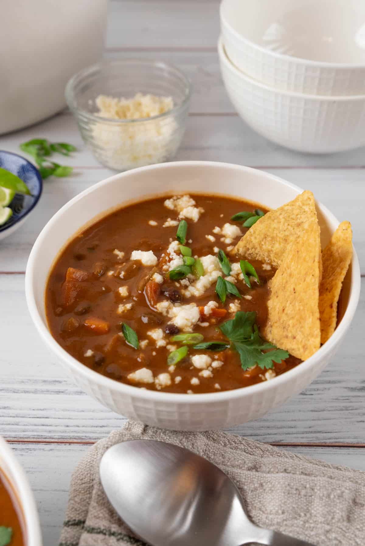A closeup of a bowl of sweet potato black bean soup garnished with queso fresco, green onions and tortilla chips, sitting with a linen napkin and a spoon. Bowls of garnishes scattered around and a stack of white bowls in the background.