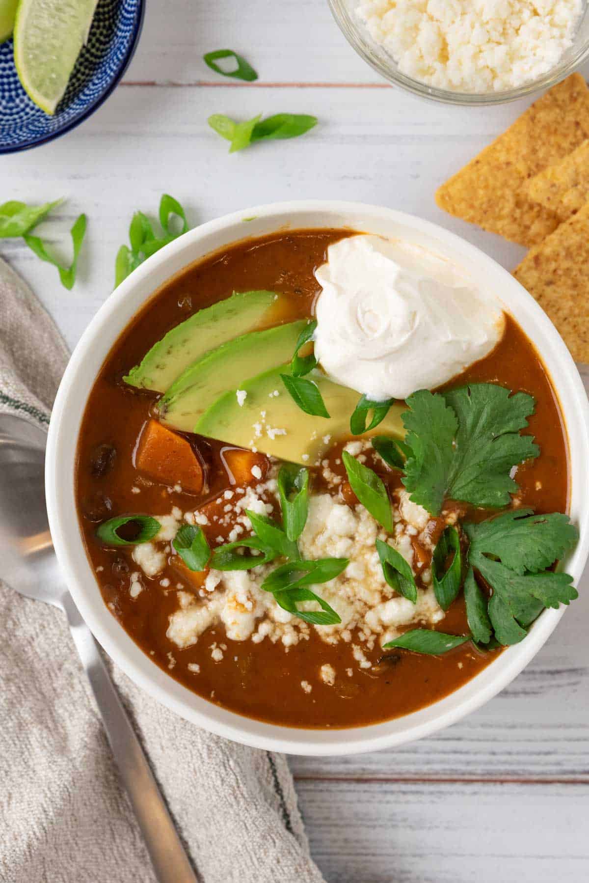 A bowl of garnished sweet potato black beans soup and a linen napkin and spoon, with a bowl of tortilla chips, a big soup pot and various bowls with garnishes.