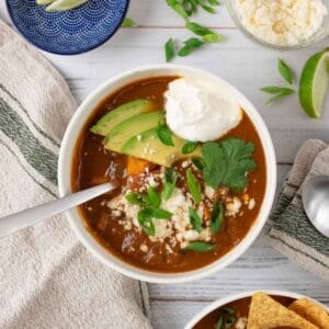 A white bowl filled with sweet potato black bean soup, garnished with queso fresco, sour cream, green onion, cilantro and avocado on a white wooden tabletop. Surrounding it is another bowl of soup, a linen napkin with a green stripe, a bowl of lime wedges, queso fresco and more sliced green onions.