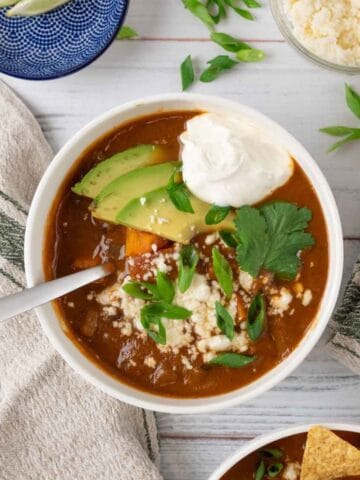 A white bowl filled with sweet potato black bean soup, garnished with queso fresco, sour cream, green onion, cilantro and avocado on a white wooden tabletop. Surrounding it is another bowl of soup, a linen napkin with a green stripe, a bowl of lime wedges, queso fresco and more sliced green onions.