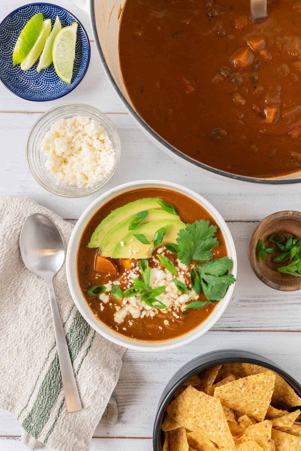 A bowl of garnished sweet potato black beans soup and a linen napkin and spoon, with a bowl of tortilla chips, a big soup pot and various bowls with garnishes.