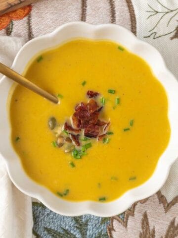 A fluted white bowl filled with butternut squash carrot soup, garnished with bacon crumbles, chives and pepitas, resting on a fall themed linen.