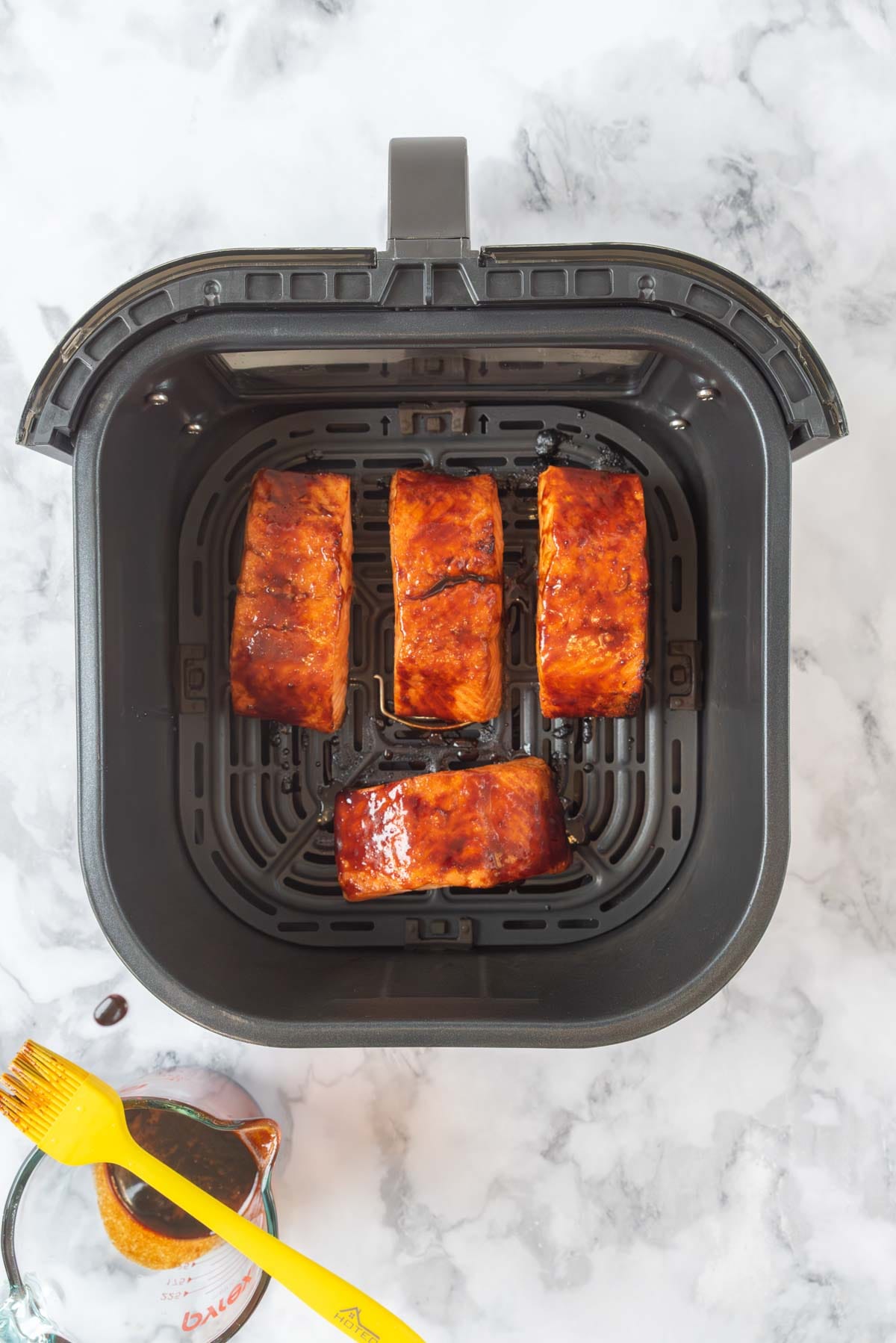 Salmon glazed in marinade sitting in an air fryer basket. A glass mixing cup of marinade and a brush sitting next to the air fryer basket.