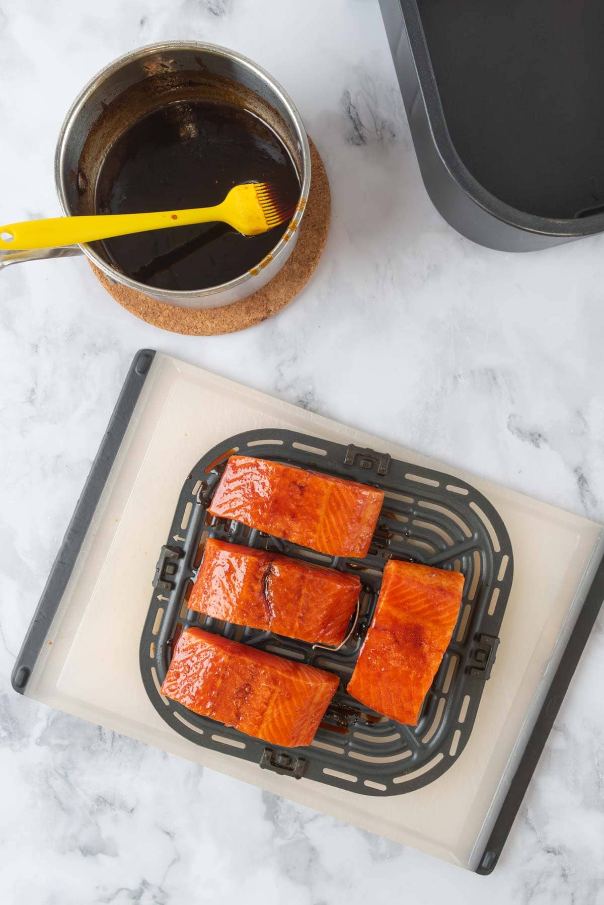 A saucepan with hot honey glaze sitting near a cutting board with an air fryer basket tray filled with salmon filets.