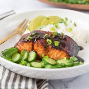 A closeup of hot honey glazed air fryer salmon plated with rice and cucumber, garnished with more glaze, chopped cilantro and green onions. A black and white striped napkin in the foreground and a wooden platter partially in frame in the background.
