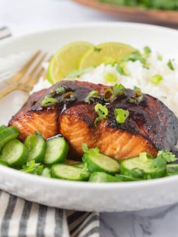 A closeup of hot honey glazed air fryer salmon plated with rice and cucumber, garnished with more glaze, chopped cilantro and green onions. A black and white striped napkin in the foreground and a wooden platter partially in frame in the background.