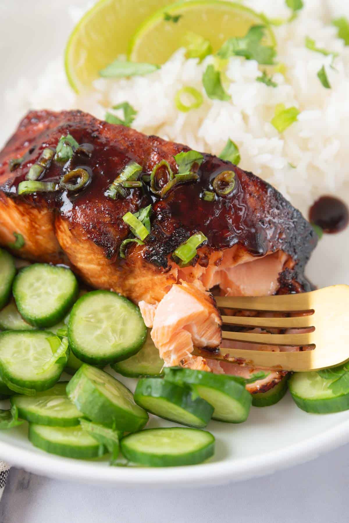 A closeup of a bite hot honey glazed air fryer salmon on a fork over the plated dish with rice and cucumber, garnished with more glaze, chopped cilantro and green onions.