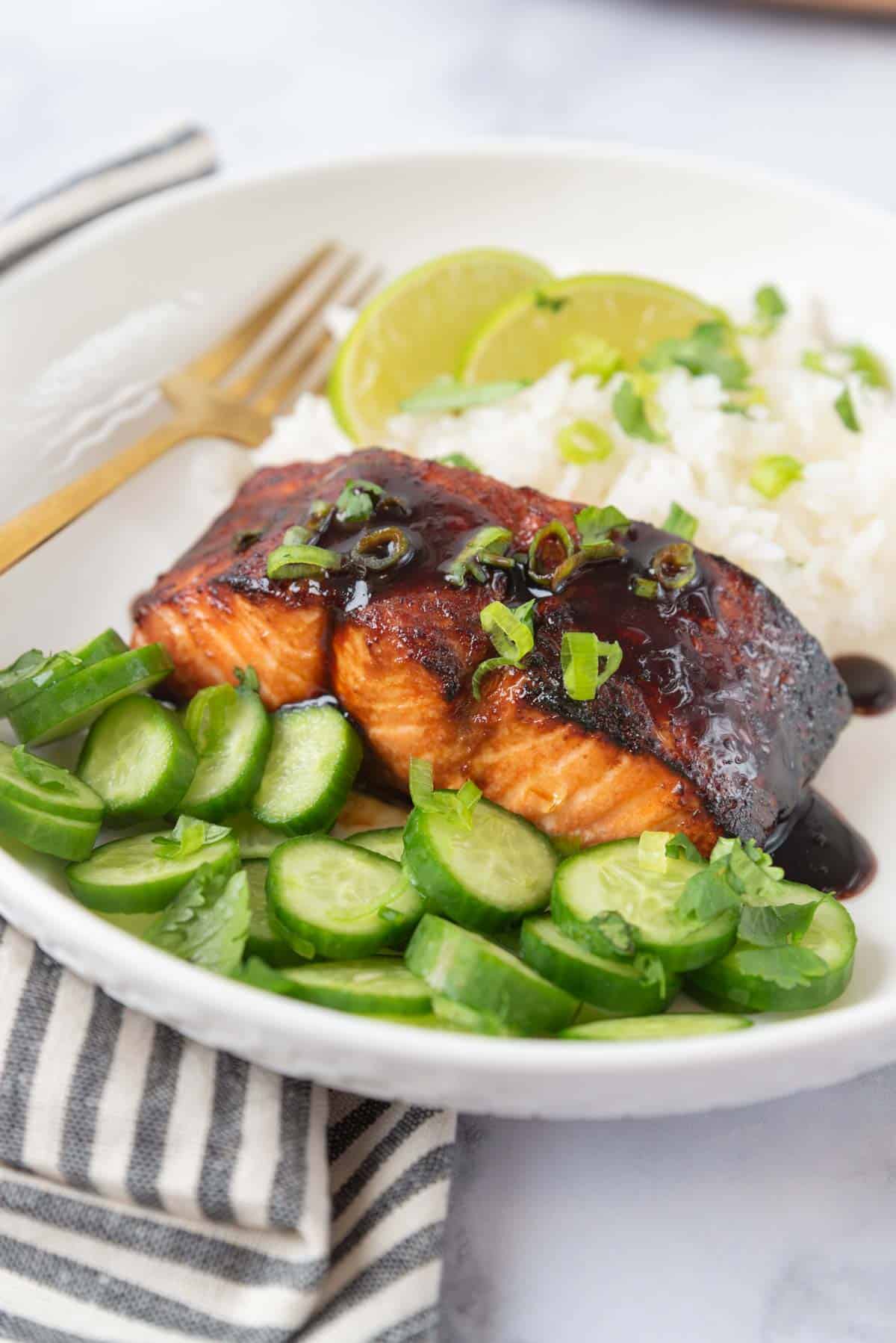 Beautifully glazed hot honey salmon plated with rice and sliced cucumber. The dish is garnished with cilantro and sliced green onions.