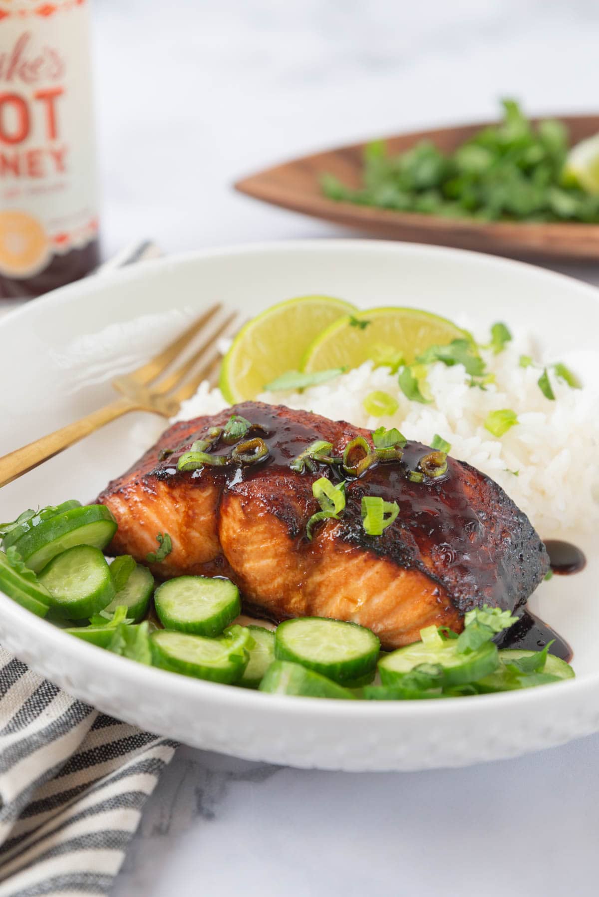 Beautifully glazed hot honey salmon plated with rice and sliced cucumber. The dish is garnished with cilantro and sliced green onions. A black and white striped linen napkin in the foreground with a bottle of hot honey and a wooden tray with more garnishes in the background.