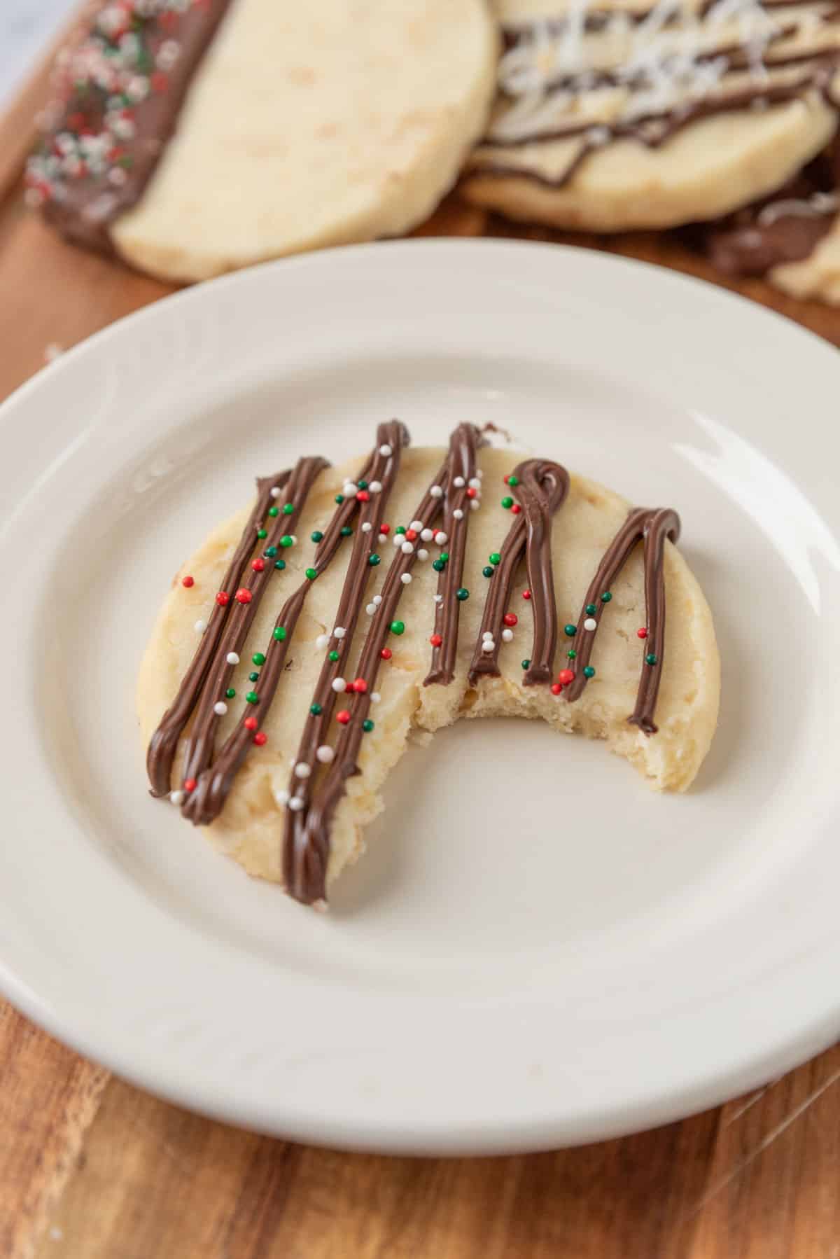 A bite taken from a cookie decorated with a chocolate drizzle and holiday sprinkles, resting on a white plate on top of a wooden tabletop.