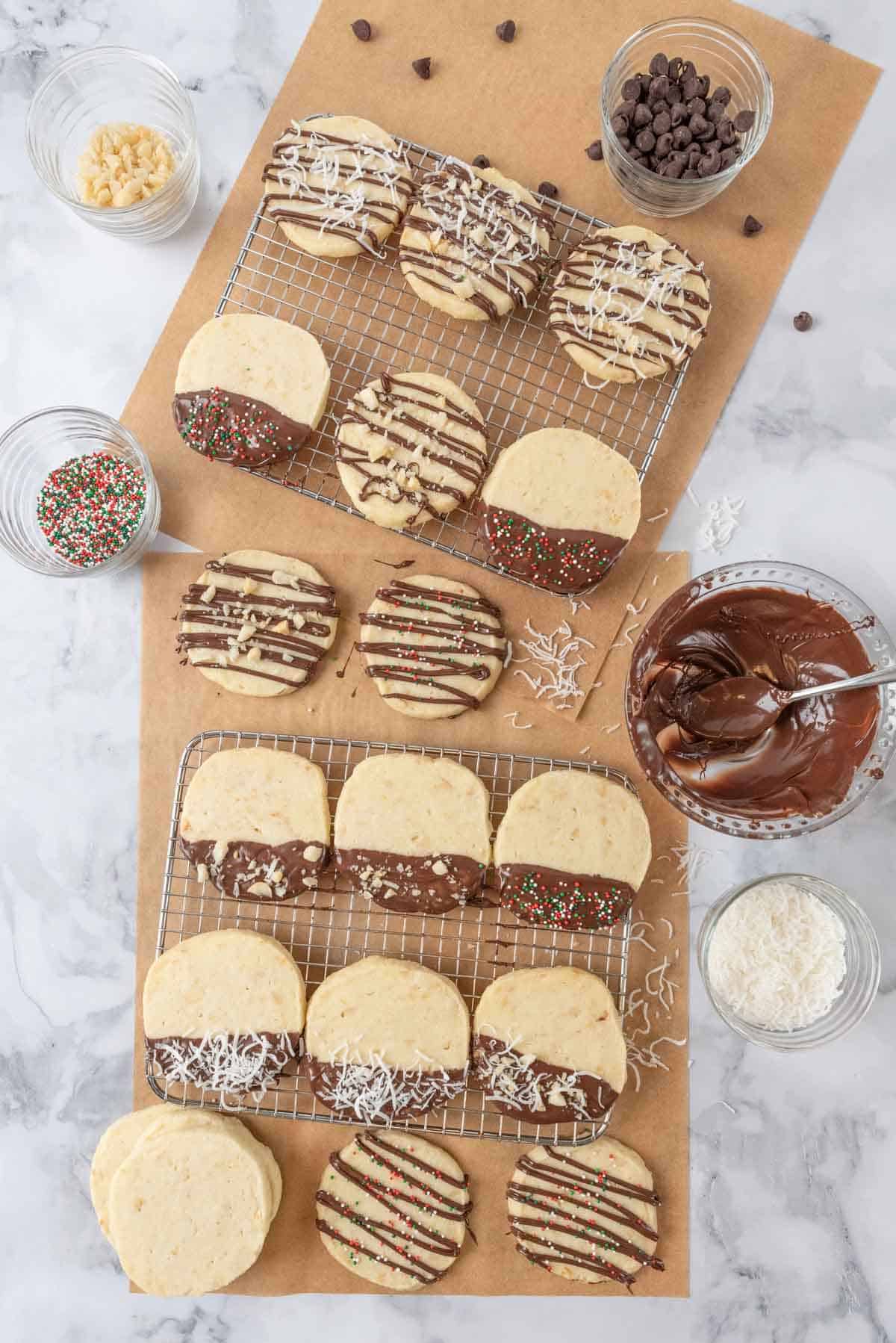 Shortbread cookies decorated on cooling racks. Small bowls of shredded coconut, chocolate chips, chopped macadamia nuts and holiday sprinkles near the cookies.