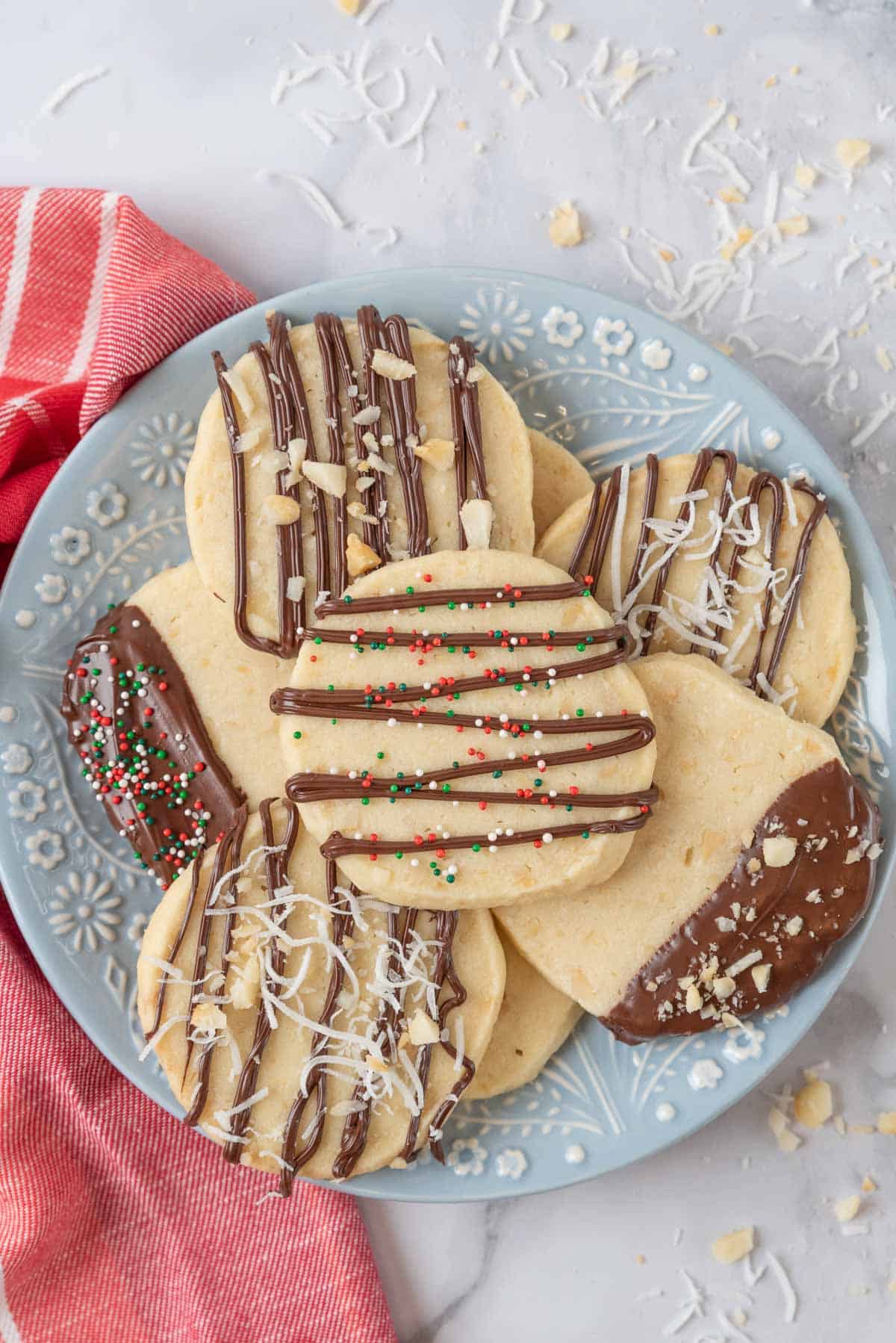 A blue and white flowered plate piled high with macadamia nut shortbread cookies. A red and white napkin sits beside the plate and chopped nuts and shredded coconut are scattered on the countertop.
