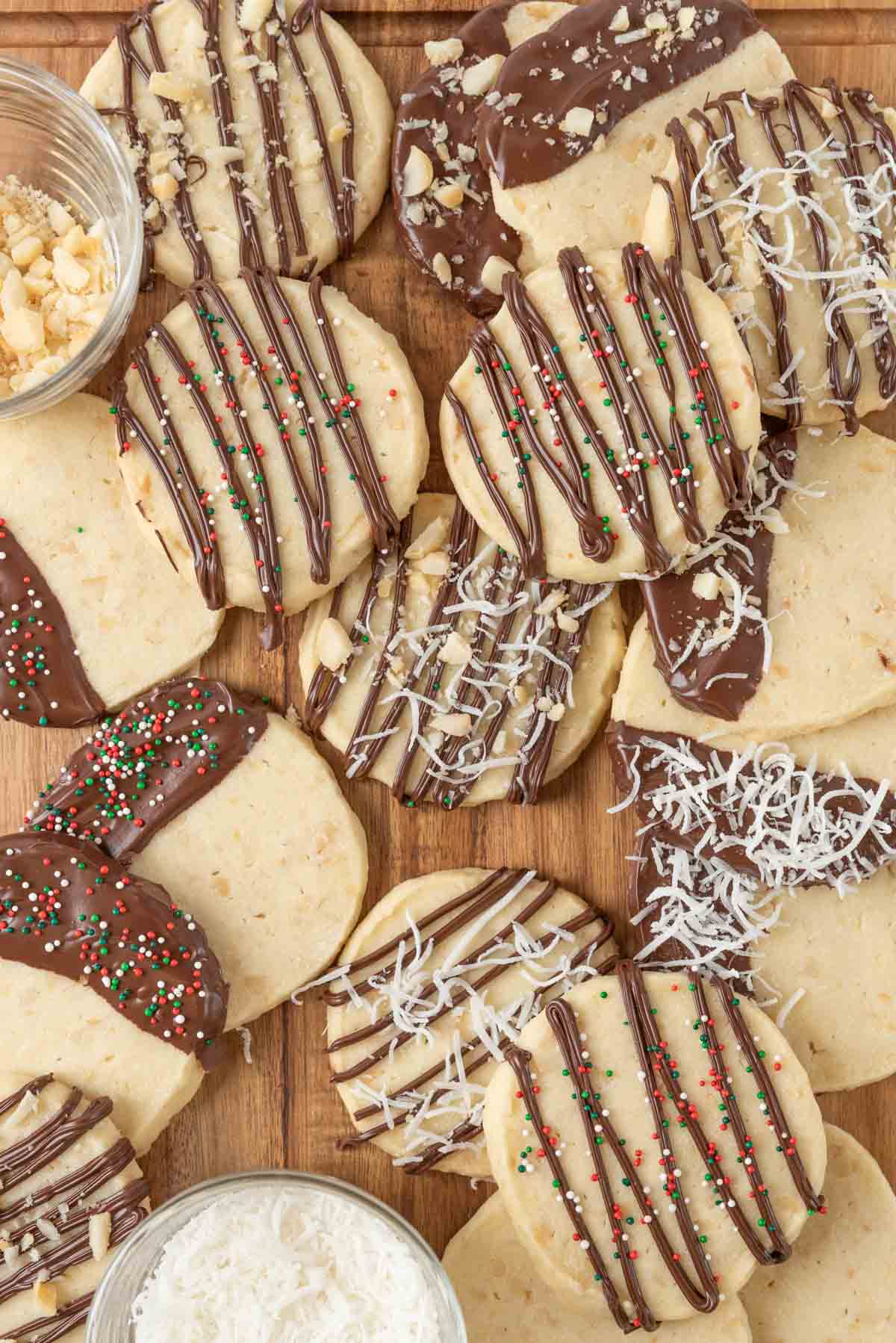 A wooden tabletop with decorated macadamia nut shortbread cookies and small bowls of shredded coconut and chopped macadamia nuts.
