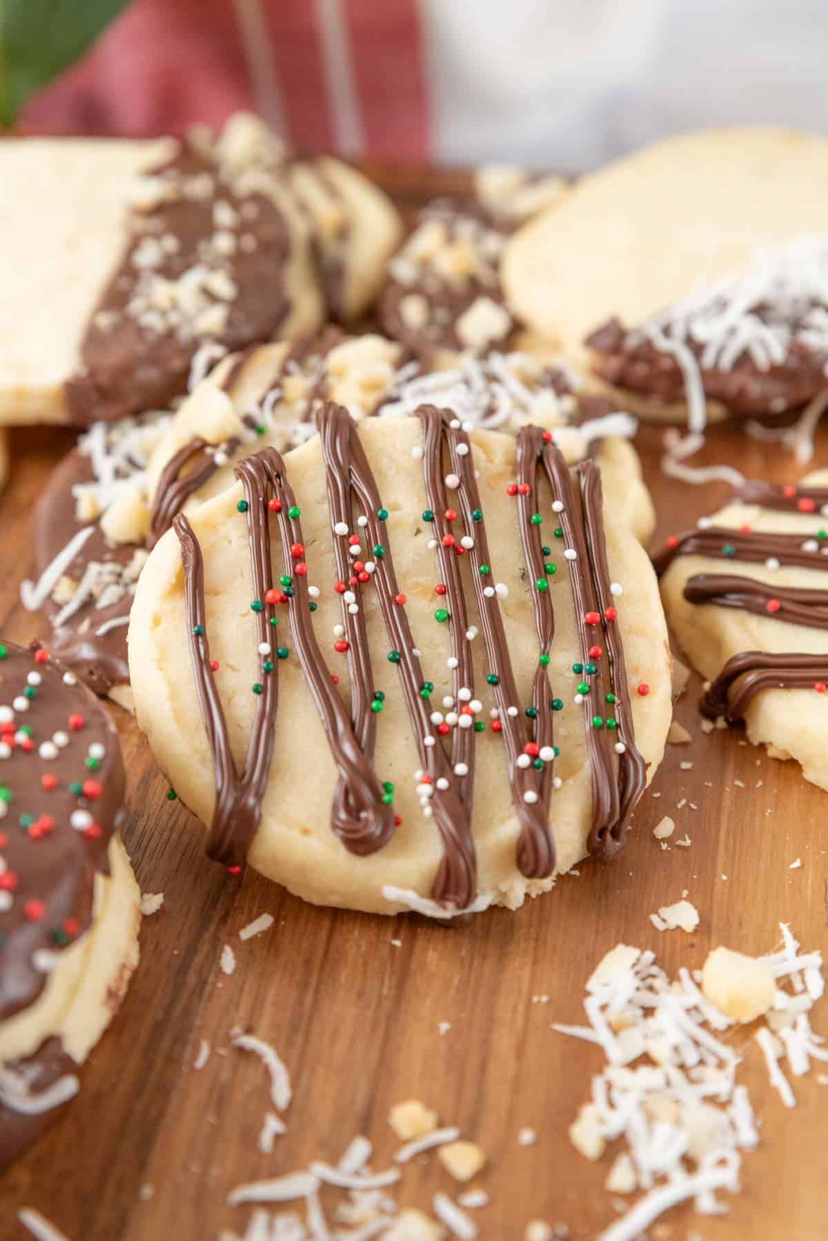 A closeup of a shortbread cookie decorated with a chocolate drizzle and holiday sprinkles. It rests on a wooden countertop scattered with more cookies and decorative toppings.