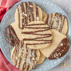 A blue and white flowered plate piled high with macadamia nut shortbread cookies. A red and white napkin sits beside the plate and chopped nuts and shredded coconut are scattered on the countertop.