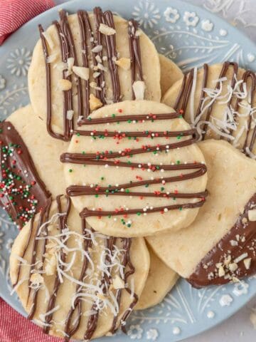 A blue and white flowered plate piled high with macadamia nut shortbread cookies. A red and white napkin sits beside the plate and chopped nuts and shredded coconut are scattered on the countertop.