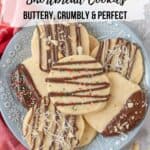 A blue and white flowered plate piled high with macadamia nut shortbread cookies. A red and white napkin sits beside the plate and chopped nuts and shredded coconut are scattered on the countertop. Graphic overlayed for Pinterest.