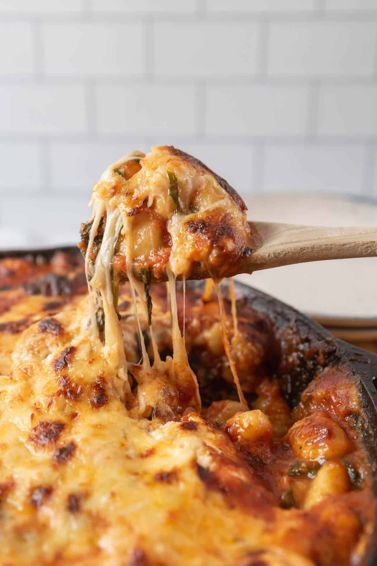 A wooden spoon filled with golden bubbly cheesy baked gnocchi, cheese stretching from the cast-iron skillet up to the spoon. Plates are in the background.