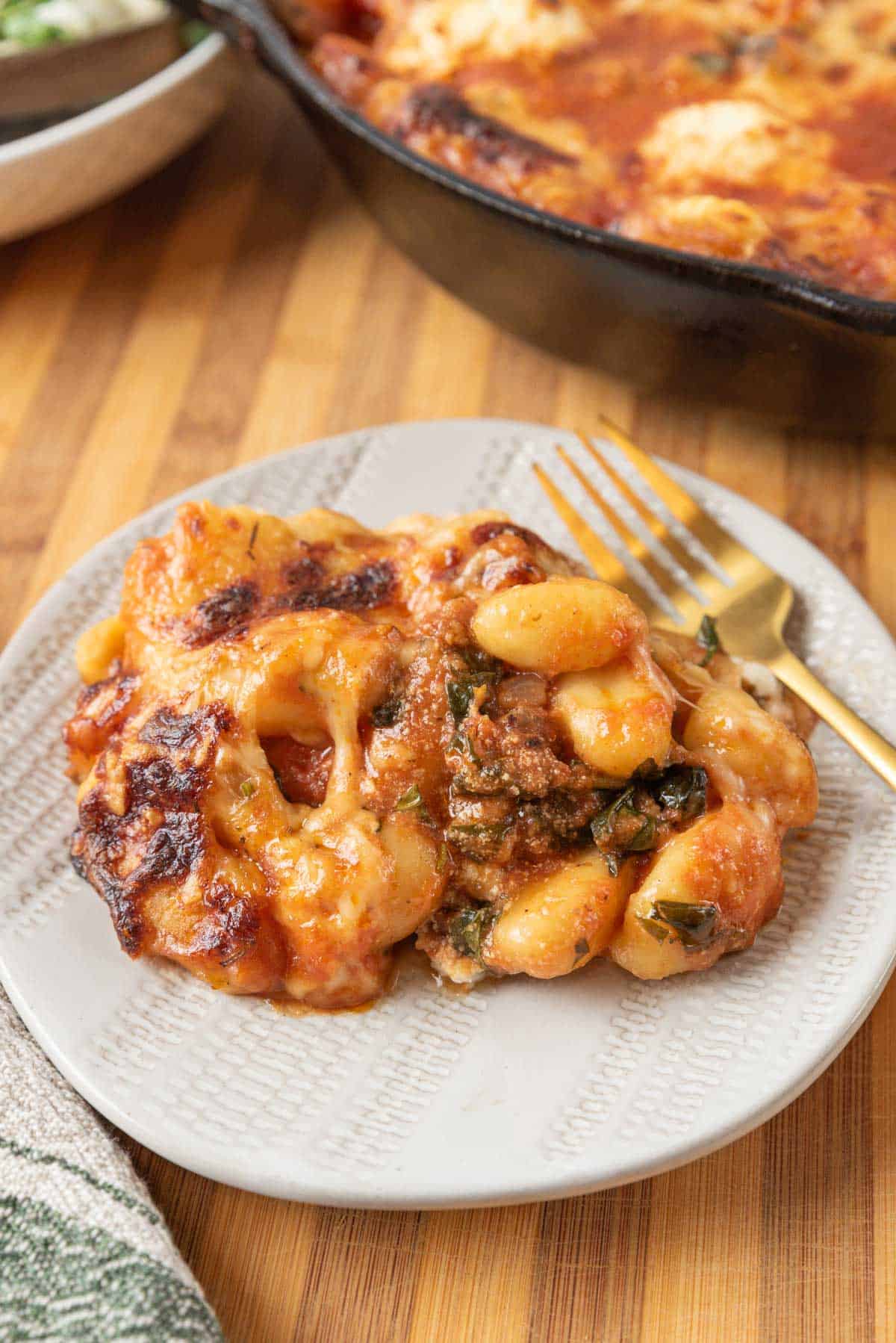 A plate of cheesy baked gnocchi with sausage and spinach with a gold fork resting on a wooden tabletop. A green salad in the background with the cast-iron skillet filled with the dish.