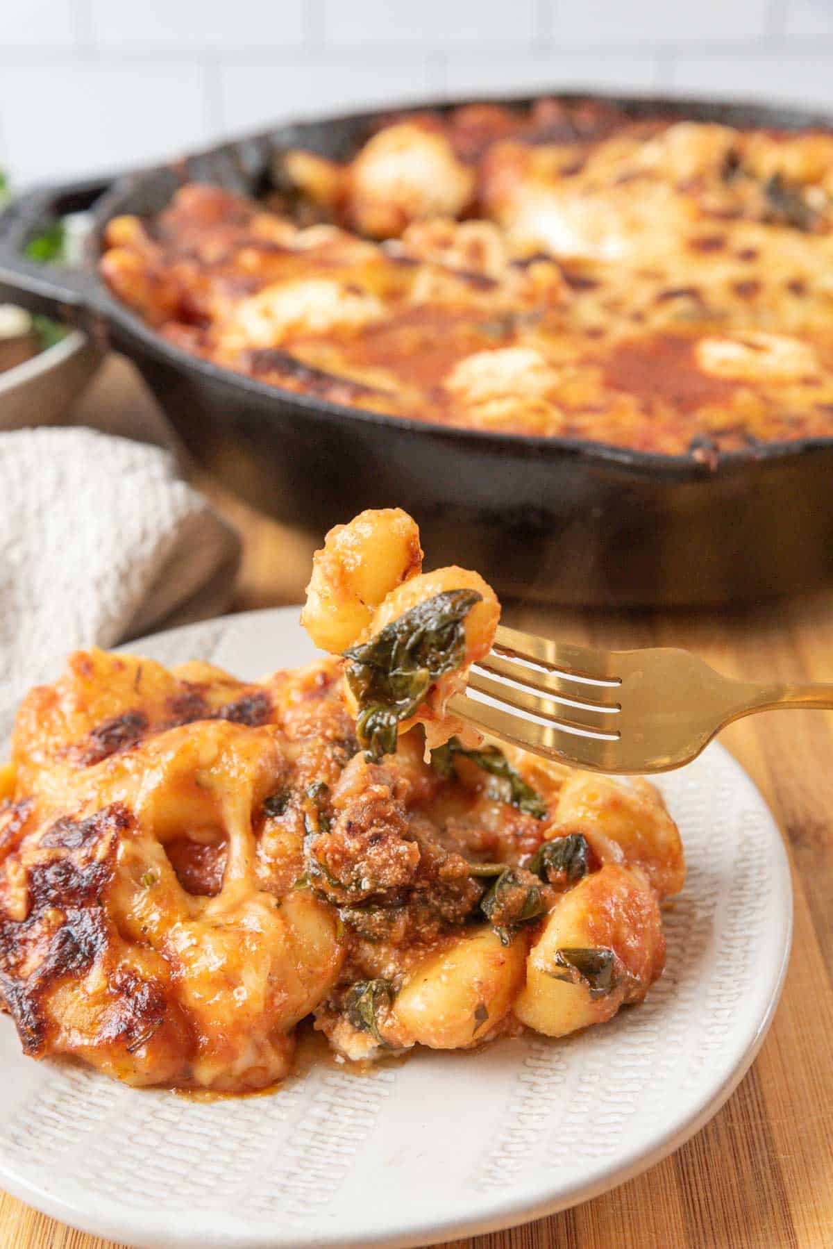 A plate of cheesy baked gnocchi with sausage and spinach with gnocchi on a gold fork, on a wooden tabletop. A green salad in the background with a linen napkin, and the cast-iron skillet filled with the pasta dish.