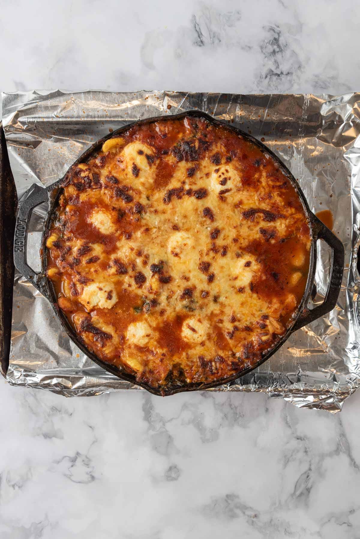 A cast-iron skillet taken from the oven with golden bubbly cheese melted on top of baked gnocchi in a cast-iron skillet. The skillet is on a baking sheet lined with aluminum foil.