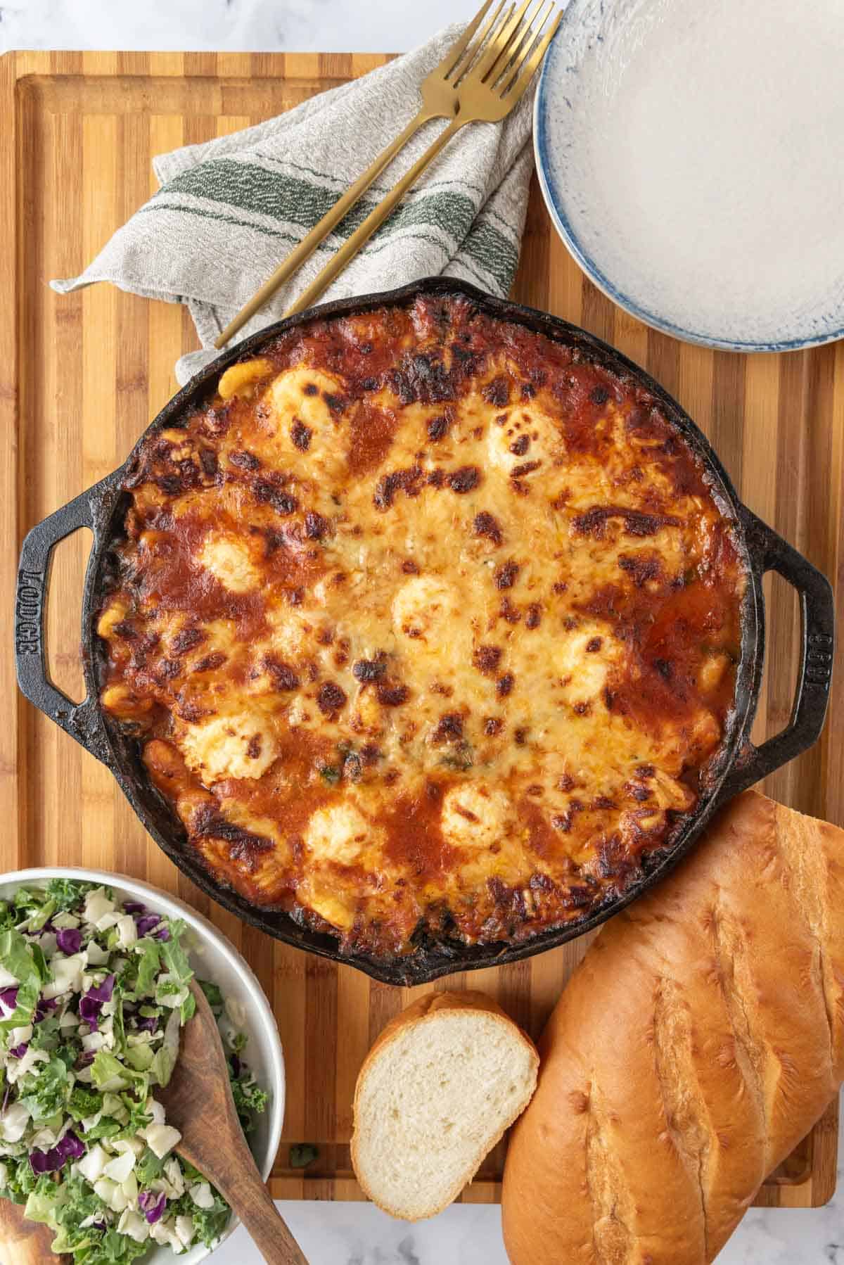 A cast-iron skillet filled with golden bubbly baked gnocchi with sausage and spinach. It's resting on a wooded surface with a bowl of salad, a loaf of sliced Italian bread, plates, napkins, and gold forks.