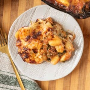 A closeup of a plate piled with golden bubbly cheesy baked gnocchi on a wooden countertop. A gold fork and linen napkin rest beside the plate and the cast-iron skillet peeks out from the corner of the frame.