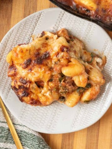 A closeup of a plate piled with golden bubbly cheesy baked gnocchi on a wooden countertop. A gold fork and linen napkin rest beside the plate and the cast-iron skillet peeks out from the corner of the frame.
