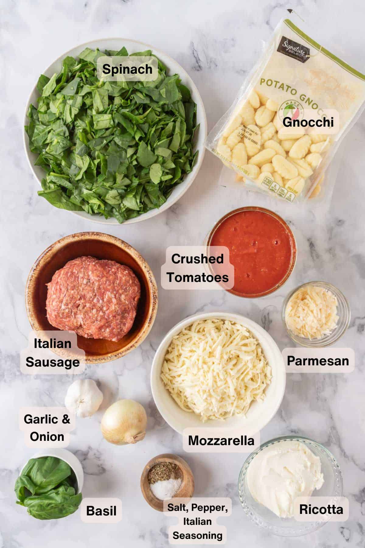 Ingredients for baked gnocchi with sausage and spinach scattered around a white marble countertop.