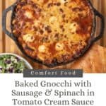 A cast-iron skillet filled with golden bubbly baked gnocchi with sausage and spinach. It's resting on a wooded surface with a bowl of salad, a loaf of Italian bread, plates, napkins, and gold forks.
