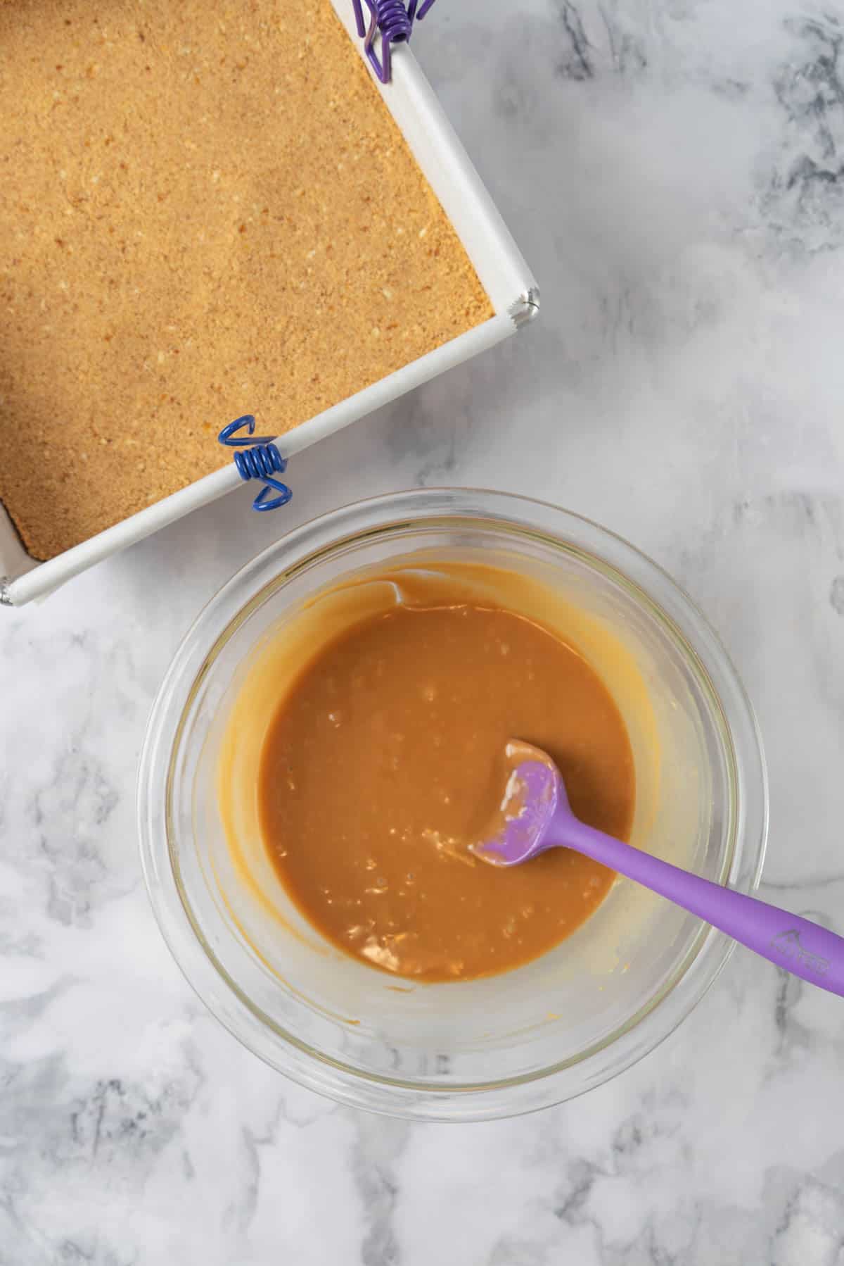 Melted caramel in a glass mixing bowl beside an 8x8 pan with the crust layer baked and set.