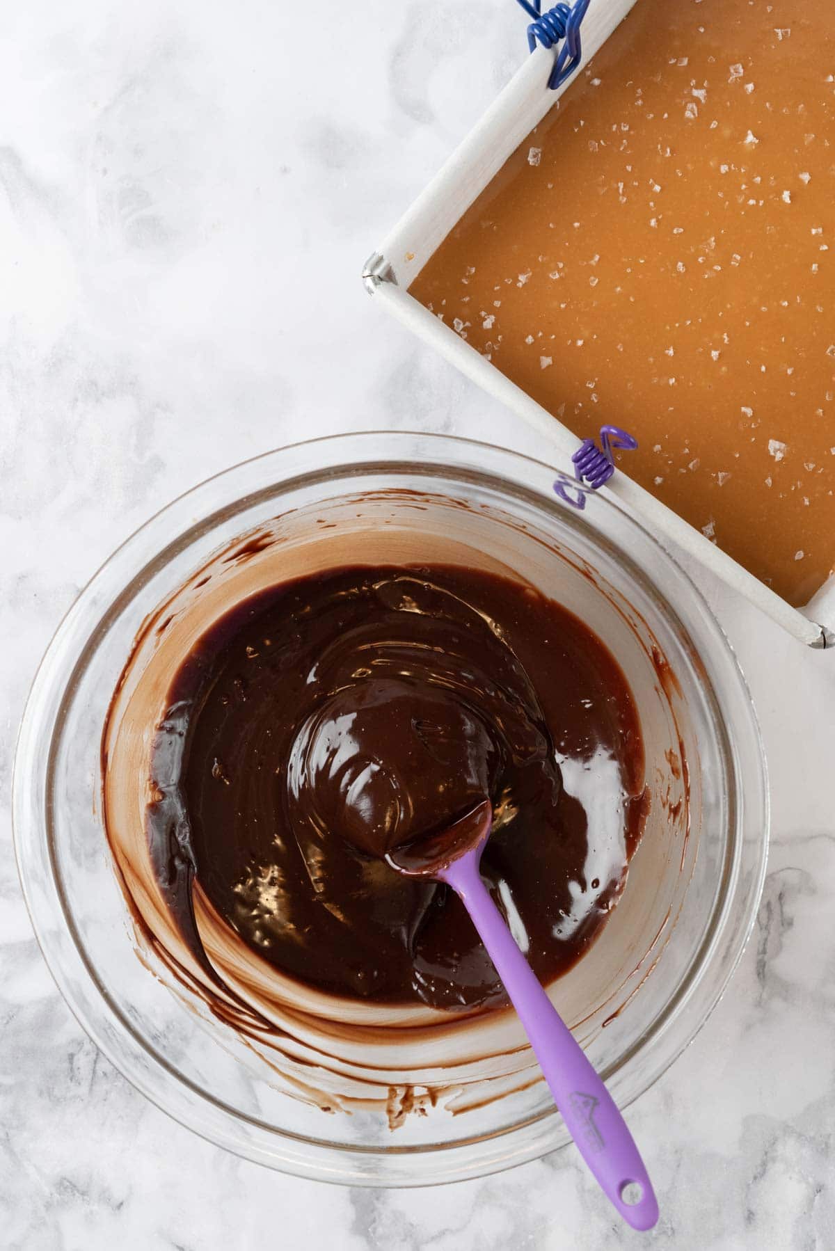 Glossy chocolate ganache in a glass mixing bowl, beside the 8x8 pan of crust and caramel layers, waiting to be poured.