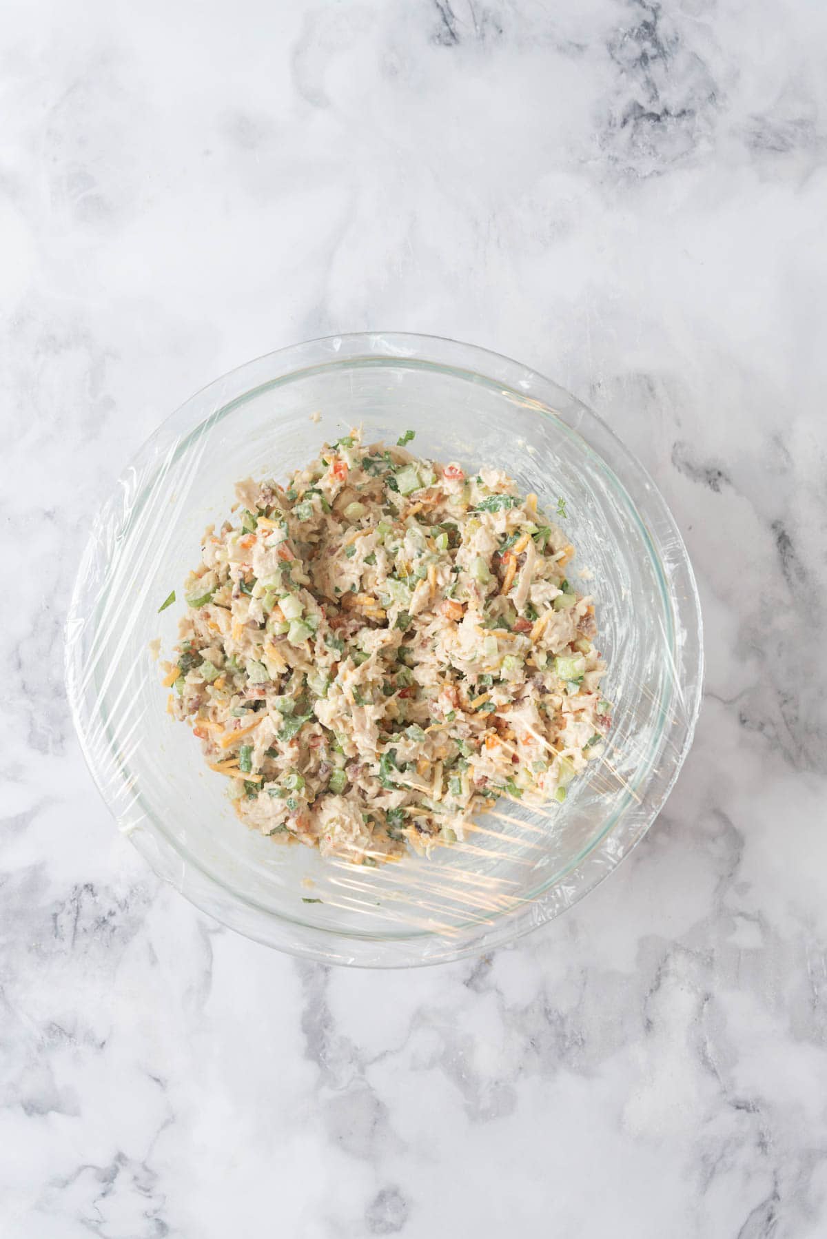 Jalapeno chicken salad mixed together in a medium glass mixing bowl with a large fork in the bowl. The bowl rests on a white and grey marble countertop, covered in plastic wrap and ready to chill in the refrigerator.