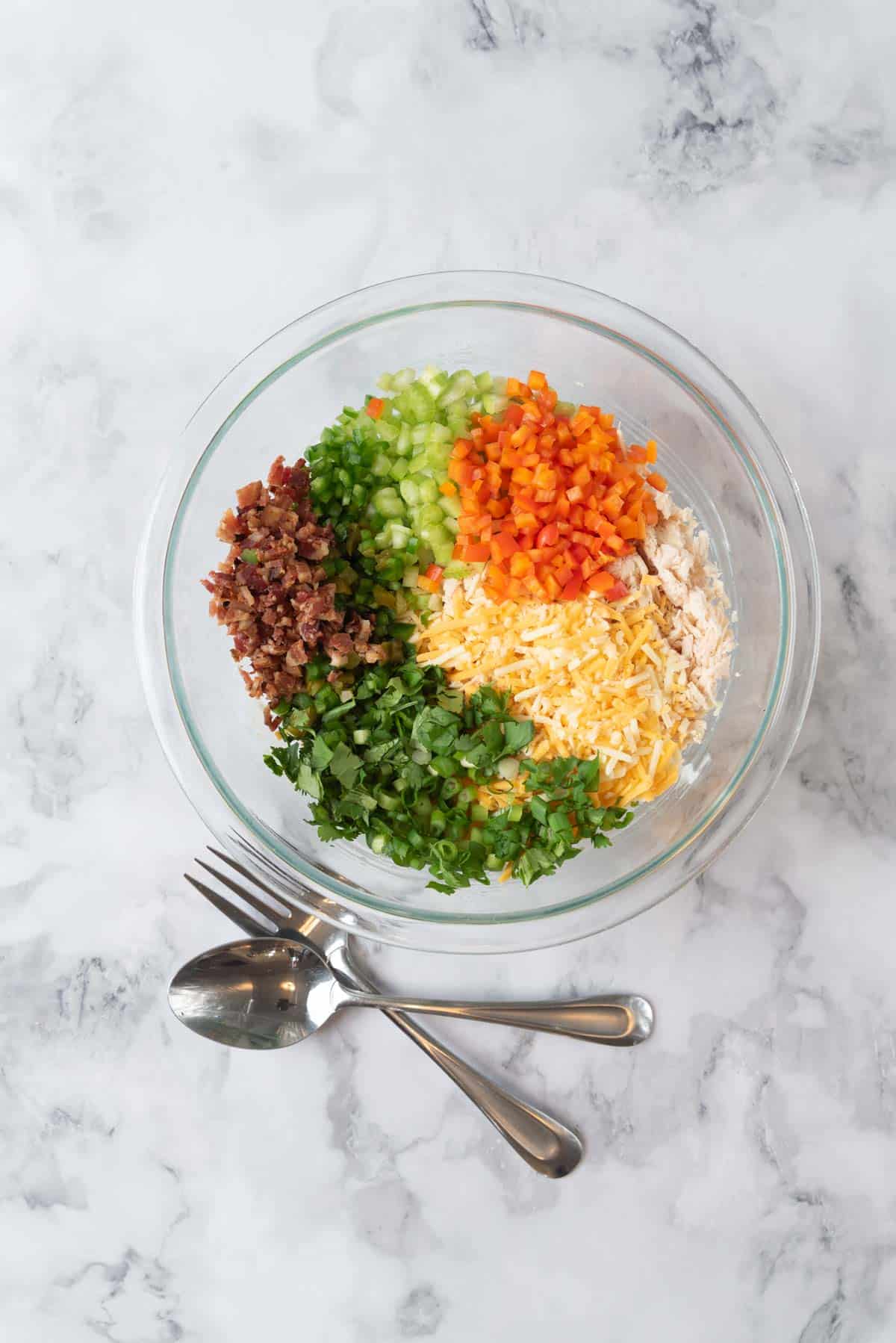 Ingredients for a jalapeno chicken salad in a medium glass mixing bowl on a white and grey marble countertop with a fork and spoon beside it, ready to be mixed together then chilled.
